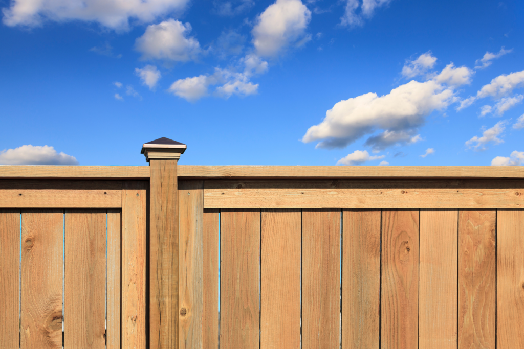 A beautifully crafted wooden fence with a decorative post cap set against a bright blue sky and fluffy white clouds, showcasing Alpine Fence Company's expert fence design and installation services in San Diego.